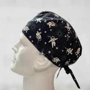 May include: A black scrub hat with a white astronaut and star pattern. The hat has a tie at the back.