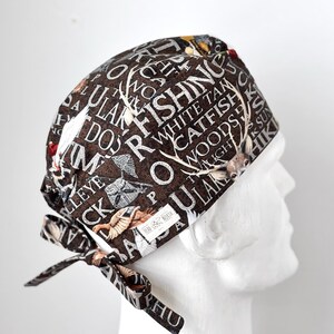 May include: A brown and white patterned surgical scrub hat with a tie. The hat features a variety of words related to fishing and hunting, including "fishing", "white tam", "catfish", "woods", and "lake".