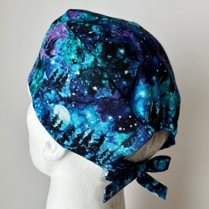 May include: A blue and purple scrub hat with a galaxy print featuring white stars and a moon. The hat has a tie at the back.