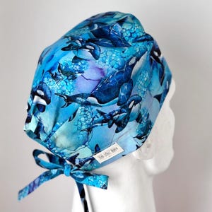 May include: Blue surgical scrub cap featuring a pattern of orca whales in shades of blue and white. The cap has a tie closure and a small tag that reads "Sea North".