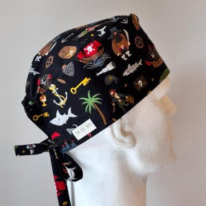 May include: Black pirate-themed scrub cap with a repeating pattern of pirate ships, treasure chests, anchors, keys, and sharks. The cap has a tie in the back and a white interior lining. The tag reads "Head North".