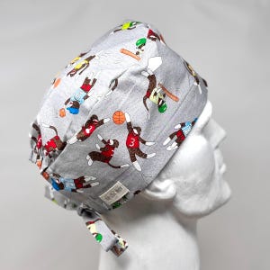 May include: A gray surgical scrub cap featuring a repeating pattern of sock monkey athletes playing basketball and baseball. The monkeys are wearing red, blue, and green uniforms. The cap is tied at the back.