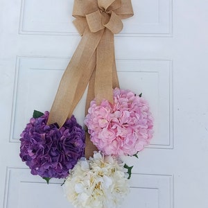 May include: A door wreath with three faux hydrangea flower heads in shades of purple, pink, and white. The wreath is tied with a burlap bow.