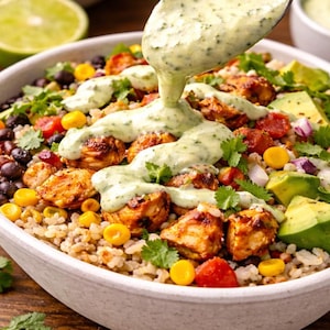 May include: A white bowl filled with a colorful salad. The salad includes grilled chicken, black beans, corn, tomatoes, avocado, and cilantro. A creamy green sauce is being poured over the salad from a spoon. A lime half is in the background.