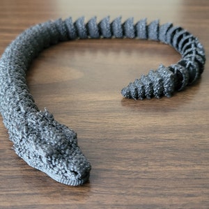 Puede incluir: Un modelo de serpiente negra impreso en 3D con una superficie texturizada. La serpiente está enroscada en forma de círculo.
