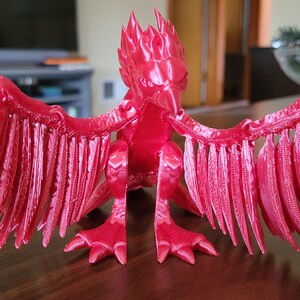 May include: A bright red 3D printed figurine of a mythical creature with wings and a fiery crest. The creature is posed in a sitting position with its wings spread out.