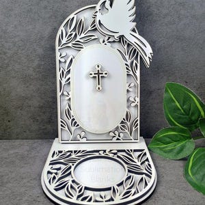 Puede incluir: Una pieza decorativa de madera blanca con una paloma, una cruz y un diseño floral. La base tiene el texto "Sublimation Blanks". La pieza está diseñada con intrincados recortes y un efecto en capas, adecuada para la sublimación.