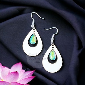 May include: A pair of white wooden teardrop earrings with iridescent teardrop charms hanging from the center. The earrings are on a black fabric background.