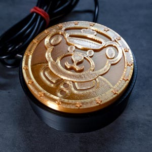 May include: A gold-colored, circular button with a bear design on top of a black base. The bear design is detailed with a hat and stars around the edge. A black cable with a red band is connected to the button.