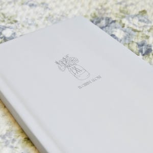 May include: A light gray journal with a floral mason jar illustration and the text "IN CHRIST ALONE" printed on the cover. The journal is lying on a floral patterned surface.