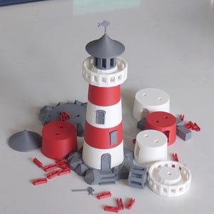 May include: A 3D-printed lighthouse model with red and white stripes, a gray top, and a small flag. The model is surrounded by various components, including gray rocks, red cylinders, and white structures. Small red clips are scattered around.