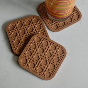 May include: Four square brown coasters with a woven rattan design. The coasters have rounded corners. One coaster has a glass with colourful stripes on top. The coasters are made of a durable material.