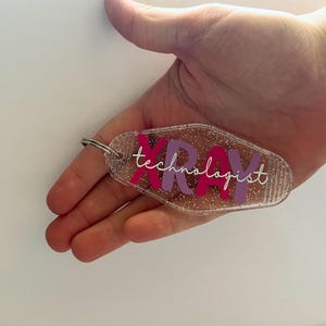 Pode incluir: Chaveiro de acrílico transparente com glitter e as palavras "XRAY technologist" em rosa e roxo. O chaveiro tem um chaveiro prateado.