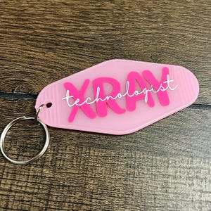 Peut inclure: Un porte-clés rose clair de style motel en forme de losange arrondi. Le mot XRAY est imprimé en grandes lettres capitales rose vif, avec le mot technologist écrit en écriture cursive blanche superposé par-dessus.