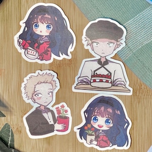 May include: A collection of anime-style stickers featuring four different characters. The stickers are on a wooden surface with green leaves and a white flower. One character holds a cake, another a pot of flowers.