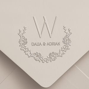 May include: A light beige envelope featuring a debossed design with the letter W centered above the text DALIA & ADRIAN. The text is framed by a symmetrical floral wreath composed of delicate leaves and small buds, creating a textured effect.
