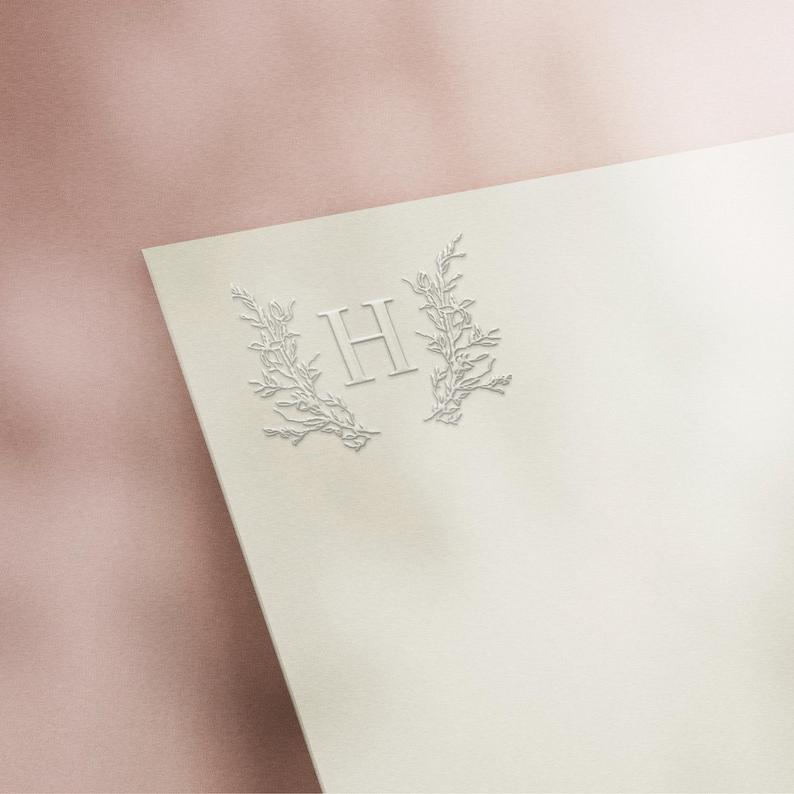 May include: Ivory-colored stationery with an embossed design. The design features the letter "H" in a serif font, framed by delicate floral branches. The paper is angled, with a soft pink background.
