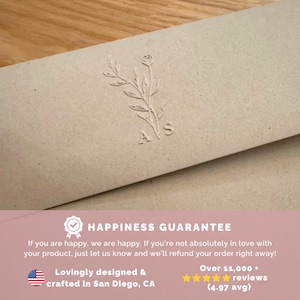 May include: A light beige paper envelope featuring a debossed botanical illustration of a leafy stem with the letters A and S positioned below. The lower section includes text stating Happiness Guarantee, refund policy, and design location details.