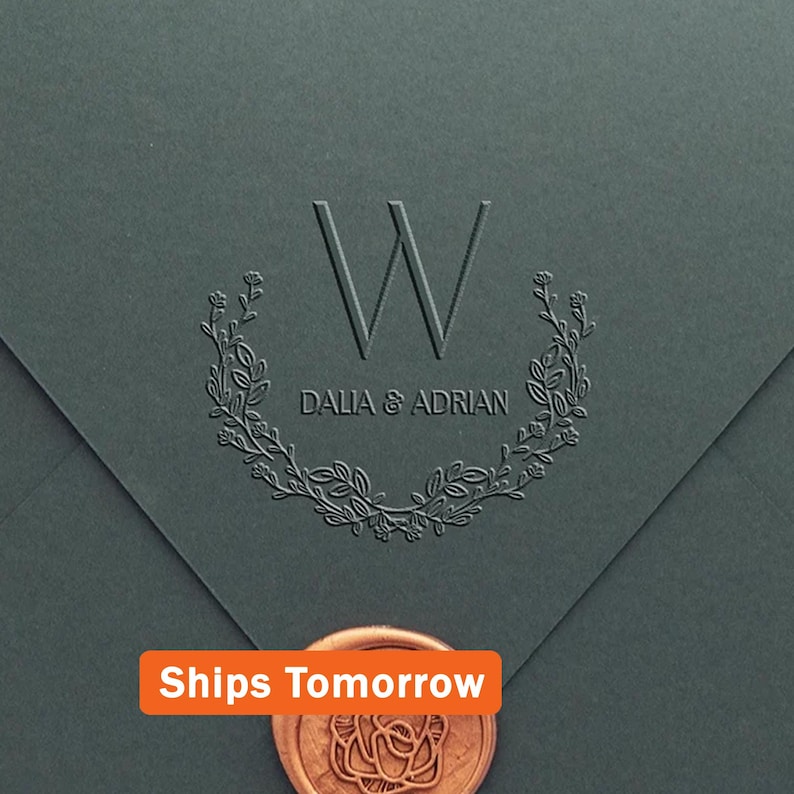 May include: A dark gray envelope featuring an embossed letter W, the names DALIA & ADRIAN, and a floral wreath design. A copper-colored wax seal with a rose pattern is positioned at the bottom, and an orange label reads Ships Tomorrow.