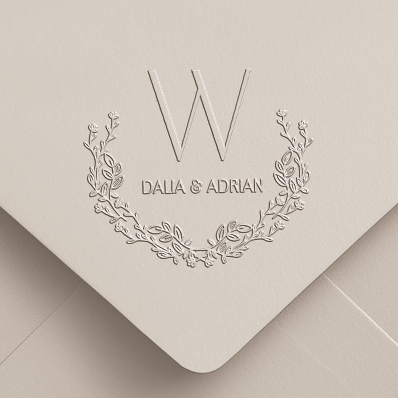 May include: A light beige envelope featuring a debossed design with the letter W centered above the text DALIA & ADRIAN. The text is framed by a symmetrical floral wreath composed of delicate leaves and small buds, creating a textured effect.