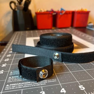 May include: Black hook and loop straps with metal snap closures. One strap is formed into a loop, and another is rolled up. The straps are on a gray cutting mat with a grid pattern. Red storage bins are in the background.
