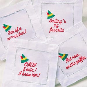 May include: Four white square coasters with embroidered text and a green, yellow, and red elf hat design. The phrases include "Son of a nutcracker!", "Smiling's my favorite", "OMG! Santa! I know him!", and "Not now, arctic puffin".