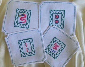 May include: Set of four white linen coasters with embroidered Mahjong tile designs. Each coaster features a different tile, with green and pink embroidery. The coasters have a scalloped edge and are arranged on a cream-colored surface.