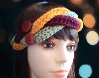 May include: A crocheted headband with a braided design in mustard yellow, burgundy, and sage green. The headband has two large, round, brown buttons.