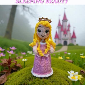 May include: A crocheted doll of Sleeping Beauty, wearing a pink dress with a white trim and a gold crown. The doll has yellow hair and is standing in a grassy area with a castle in the background. The text "SLEEPING BEAUTY" is above the doll.