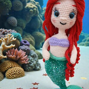 May include: Crocheted mermaid doll with red hair, a purple top, and a green sequined tail. The doll is standing on a sandy surface with orange starfish and coral in the background. The doll is approximately 10 inches tall.