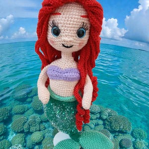 May include: A handmade crochet mermaid doll with red hair, blue eyes, and a purple top. The mermaid has a green sequined tail and is posed in a tropical ocean setting with coral and blue water.
