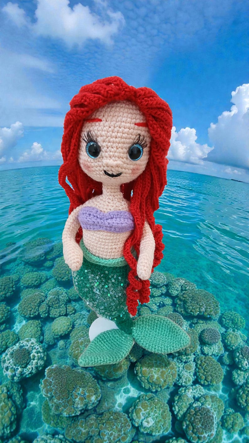 May include: A handmade crochet mermaid doll with red hair, blue eyes, and a purple top. The mermaid has a green sequined tail and is posed in a tropical ocean setting with coral and blue water.