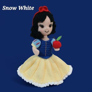 May include: Crocheted Snow White doll with a red bow, black hair, and a blue and yellow dress. The doll is holding a red apple with a green leaf. The text "Snow White" is at the top left.