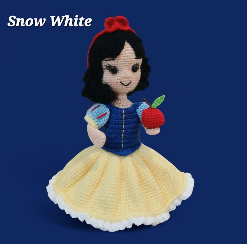 May include: Crocheted Snow White doll with a red bow, black hair, and a blue and yellow dress. The doll is holding a red apple with a green leaf. The text "Snow White" is at the top left.