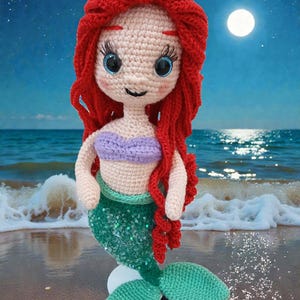 May include: A crocheted mermaid doll with red hair, blue eyes, and a purple top. The doll has a green sequined tail and is posed on a sandy beach with a full moon and starry sky in the background.