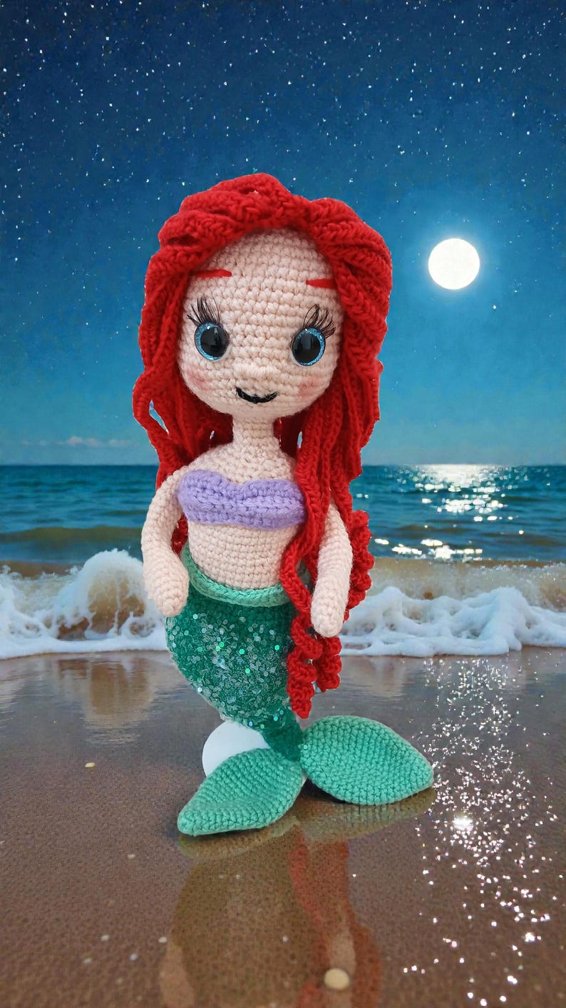 May include: A crocheted mermaid doll with red hair, blue eyes, and a purple top. The doll has a green sequined tail and is posed on a sandy beach with a full moon and starry sky in the background.
