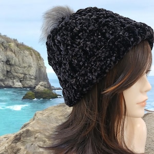 May include: A black beanie with a textured, curly surface and a fluffy gray pom-pom on top. The hat features a folded brim and is worn on a mannequin head with long, straight brown hair that has subtle reddish highlights.