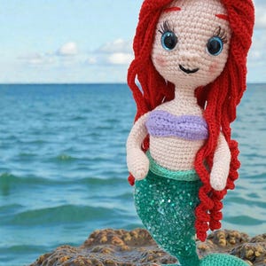 May include: A handmade crochet mermaid doll with red hair, a purple top, and a green sequined tail. The doll has blue eyes and is posed against a backdrop of the ocean and sky. The doll is approximately 10 inches tall.