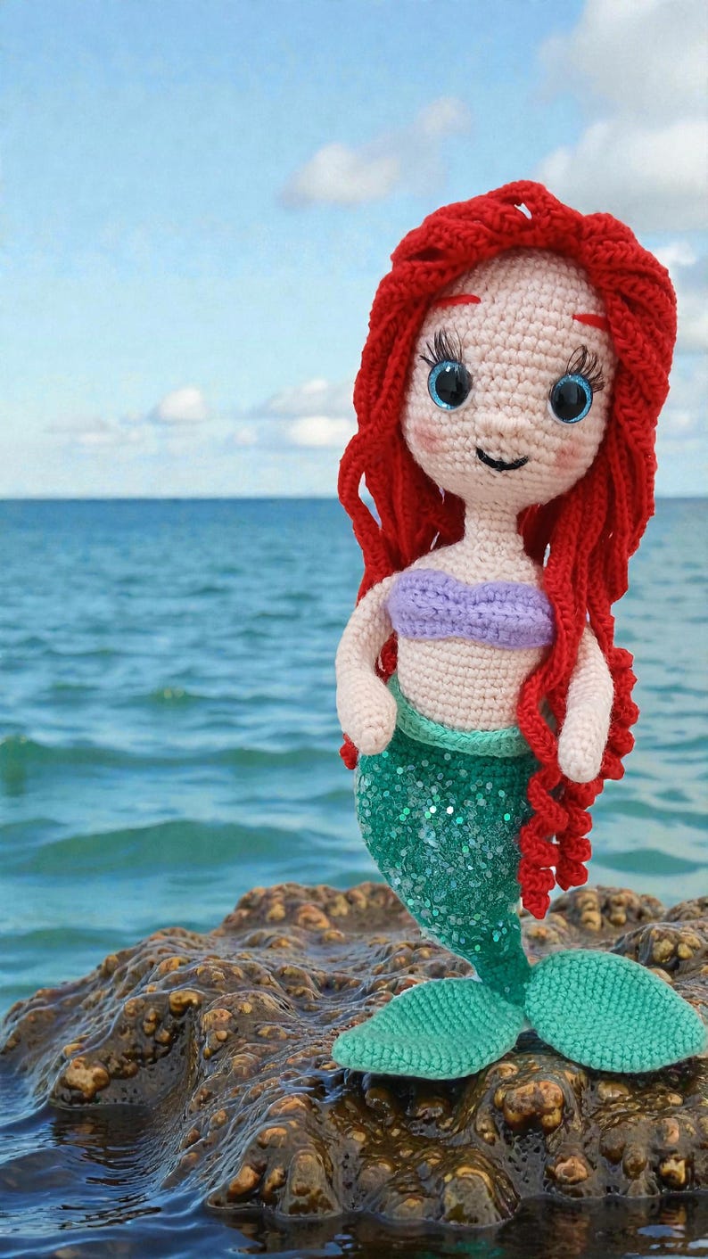 May include: A handmade crochet mermaid doll with red hair, a purple top, and a green sequined tail. The doll has blue eyes and is posed against a backdrop of the ocean and sky. The doll is approximately 10 inches tall.