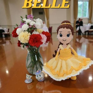 May include: A crocheted doll in a yellow dress, inspired by Belle, with brown hair and a yellow headband. The word "BELLE" is displayed in yellow text. A vase of flowers and two small figurines are also present.