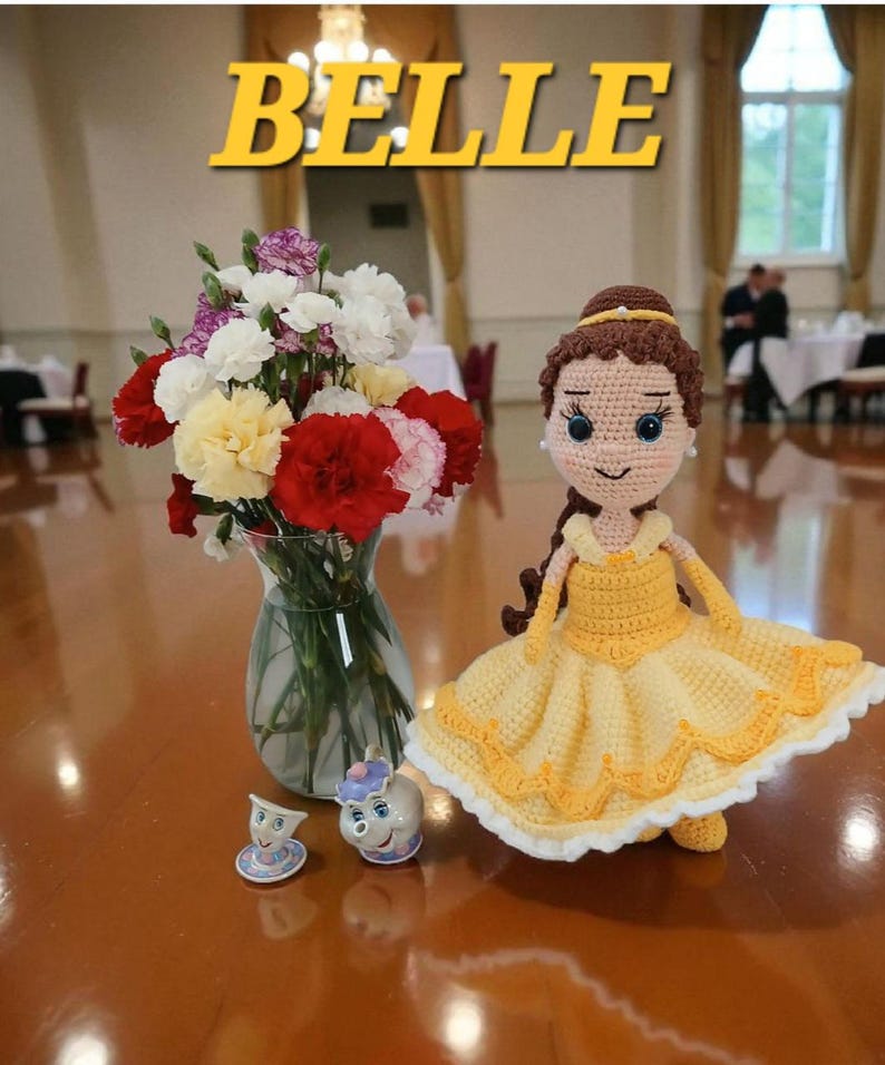 May include: A crocheted doll in a yellow dress, inspired by Belle, with brown hair and a yellow headband. The word "BELLE" is displayed in yellow text. A vase of flowers and two small figurines are also present.