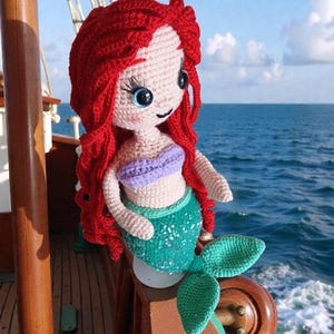 May include: Crocheted mermaid doll with red hair, blue eyes, and a green sequined tail. The doll wears a purple top and is positioned on a wooden boat railing with the ocean in the background. A handmade, whimsical item.
