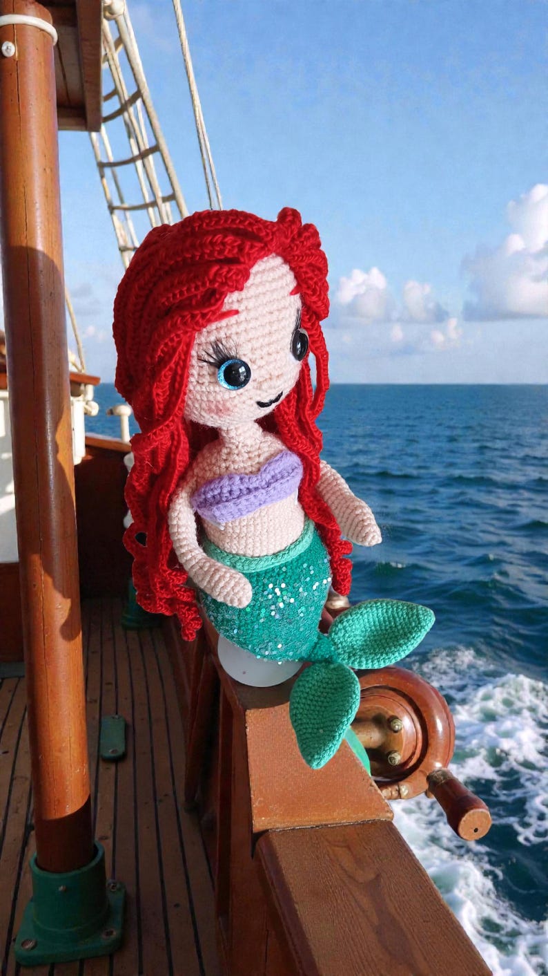 May include: Crocheted mermaid doll with red hair, blue eyes, and a green sequined tail. The doll wears a purple top and is positioned on a wooden boat railing with the ocean in the background. A handmade, whimsical item.