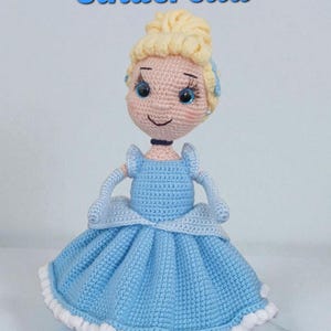 May include: A handmade crochet doll of Cinderella, featuring a light blue dress with a white trim. The doll has blonde hair styled in a bun, blue eyes, and the word "Cinderella" in blue script above.