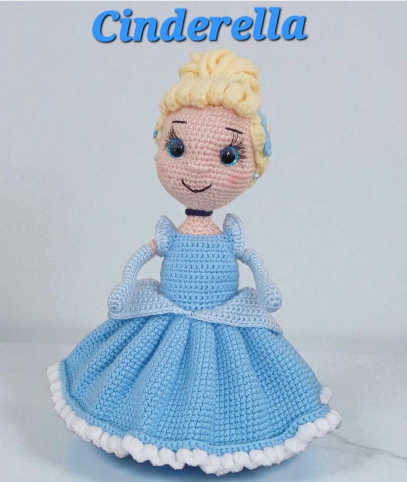 May include: A handmade crochet doll of Cinderella, featuring a light blue dress with a white trim. The doll has blonde hair styled in a bun, blue eyes, and the word "Cinderella" in blue script above.