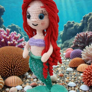 May include: A handmade crochet mermaid doll with red hair, a purple top, and a green sequined tail. The doll is standing on a bed of pebbles and seashells, with a colorful coral reef background.