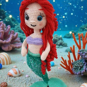 May include: A handmade crochet mermaid doll with red hair, blue eyes, and a purple top. The mermaid has a green sequined tail and is posed on a sandy surface with seashells and coral in the background. The doll is approximately 10 inches tall.