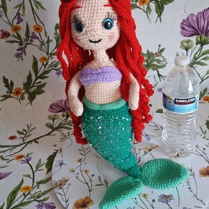 May include: Crocheted mermaid doll with red hair, a purple top, and a green sequined tail. The doll has blue eyes and is displayed on a floral patterned surface. A water bottle is next to the doll.
