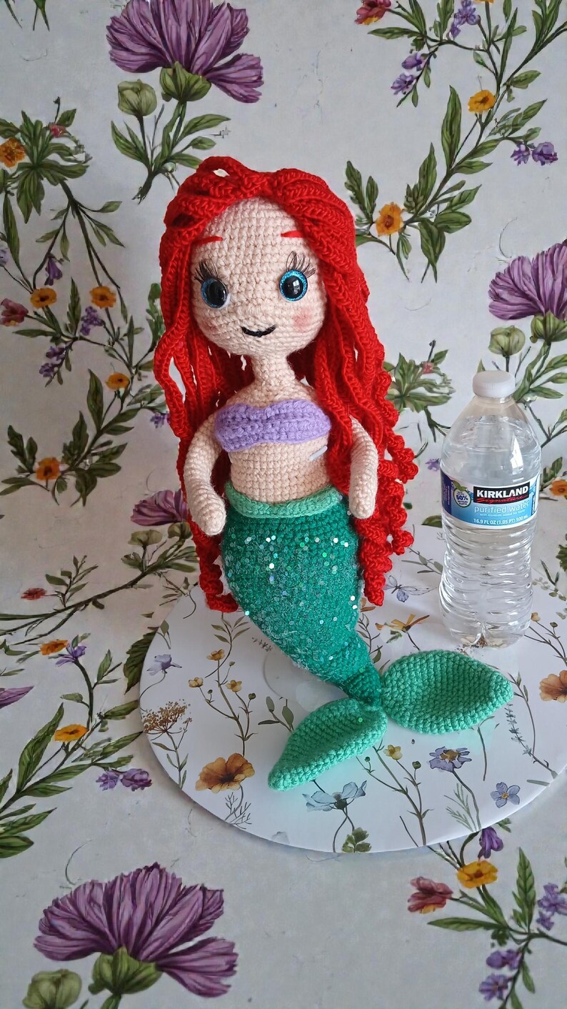 May include: Crocheted mermaid doll with red hair, a purple top, and a green sequined tail. The doll has blue eyes and is displayed on a floral patterned surface. A water bottle is next to the doll.