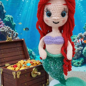 May include: Crocheted mermaid doll with red hair, a purple top, and a green sequined tail. The doll is set against an underwater backdrop with a treasure chest filled with gold coins and colorful gems. Fish swim in the background.
